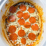 A white slow cooker containing a layer of melted cheese and pepperoni slices over a cooked, saucy base, resembling a pizza dip. The sides of the cooker are stained with sauce.
