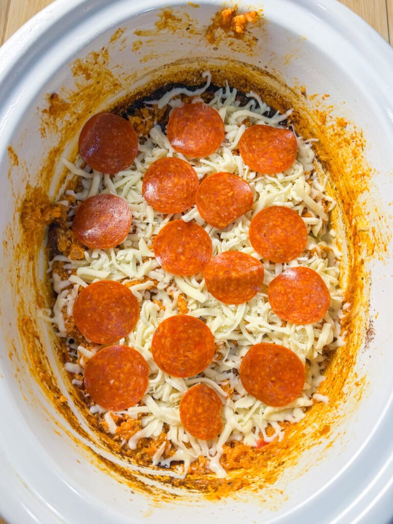 A white slow cooker containing a layer of melted cheese and pepperoni slices over a cooked, saucy base, resembling a pizza dip. The sides of the cooker are stained with sauce.