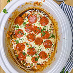Crockpot filled with a baked dish topped with melted cheese, sliced pepperoni, and chopped parsley. A small bowl of shredded cheese and scattered parsley are nearby on a striped cloth and wooden surface.
