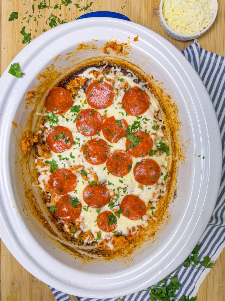 Crockpot filled with a baked dish topped with melted cheese, sliced pepperoni, and chopped parsley. A small bowl of shredded cheese and scattered parsley are nearby on a striped cloth and wooden surface.