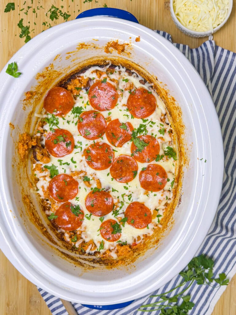 A crockpot filled with a baked dish topped with melted cheese, pepperoni slices, and chopped parsley sits on a striped cloth. A bowl of shredded cheese and fresh parsley are nearby on a wooden surface.
