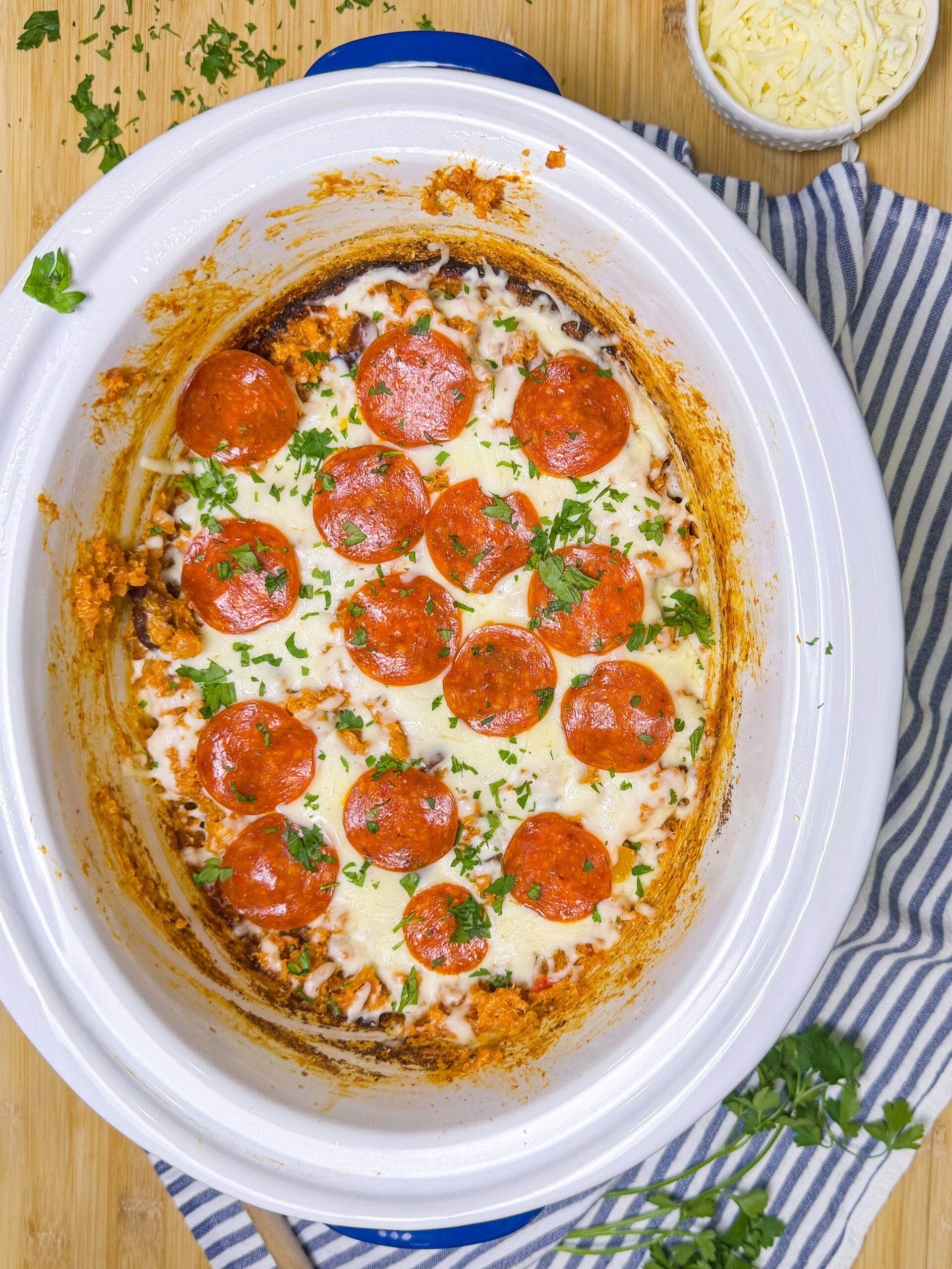 A crockpot filled with a baked dish topped with melted cheese, pepperoni slices, and chopped parsley sits on a striped cloth. A bowl of shredded cheese and fresh parsley are nearby on a wooden surface.