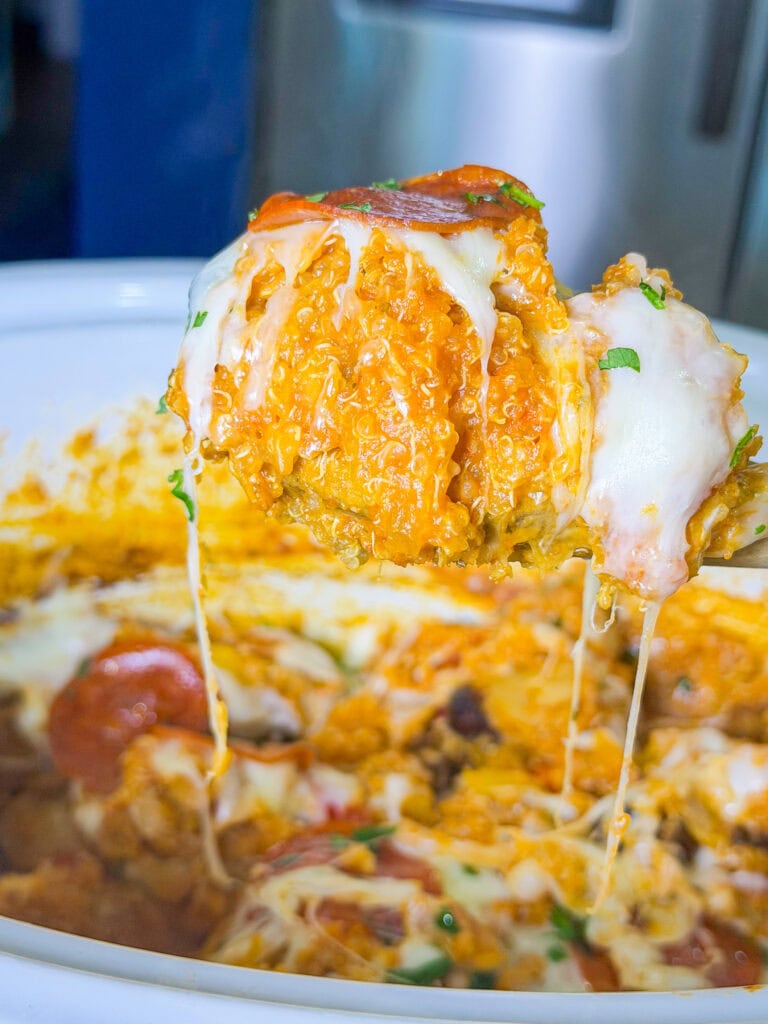 A cheesy, saucy serving of casserole is being lifted from a white dish, with melted cheese stretching and visible toppings like pepperoni and herbs.