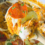 A close-up of a spoonful of cheesy, baked quinoa casserole topped with a slice of pepperoni and chopped herbs, being lifted from a dish filled with melted cheese and sauce.