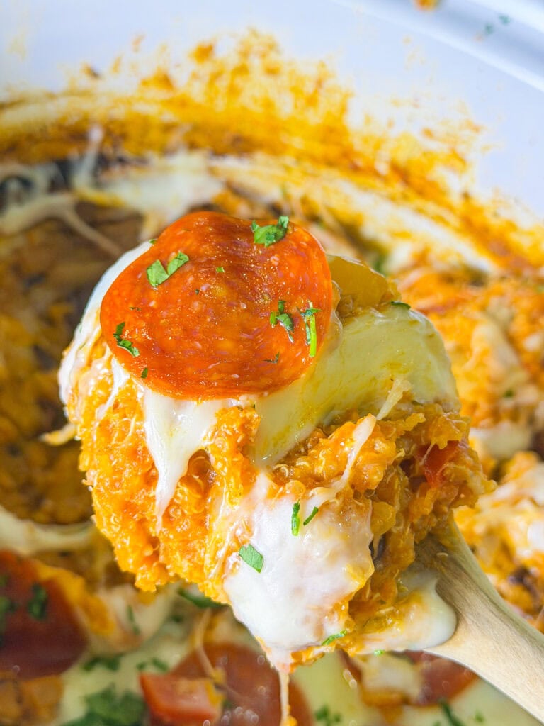 A close-up of a spoonful of cheesy, baked quinoa casserole topped with a slice of pepperoni and chopped herbs, being lifted from a dish filled with melted cheese and sauce.