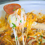 A wooden spoon lifts a cheesy, gooey serving of a casserole with melted mozzarella, pepperoni slices, and fresh herbs from a white dish.