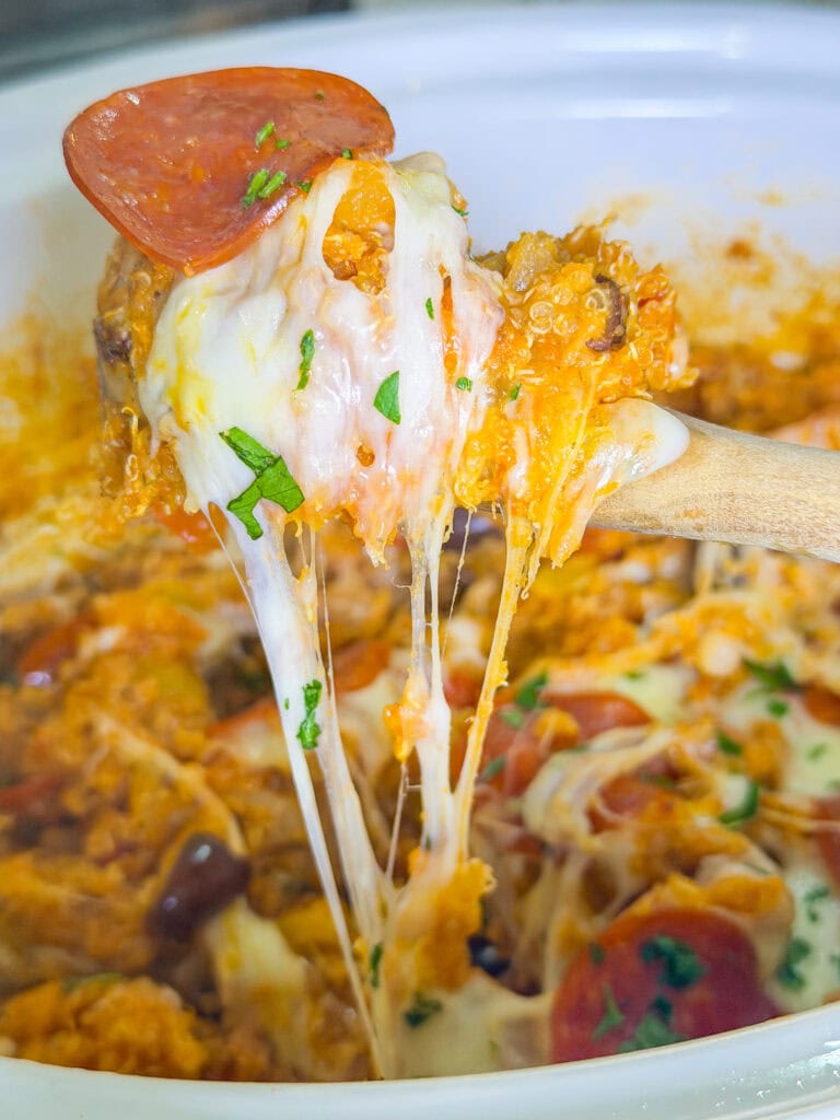 A wooden spoon lifts a cheesy, gooey serving of a casserole with melted mozzarella, pepperoni slices, and fresh herbs from a white dish.