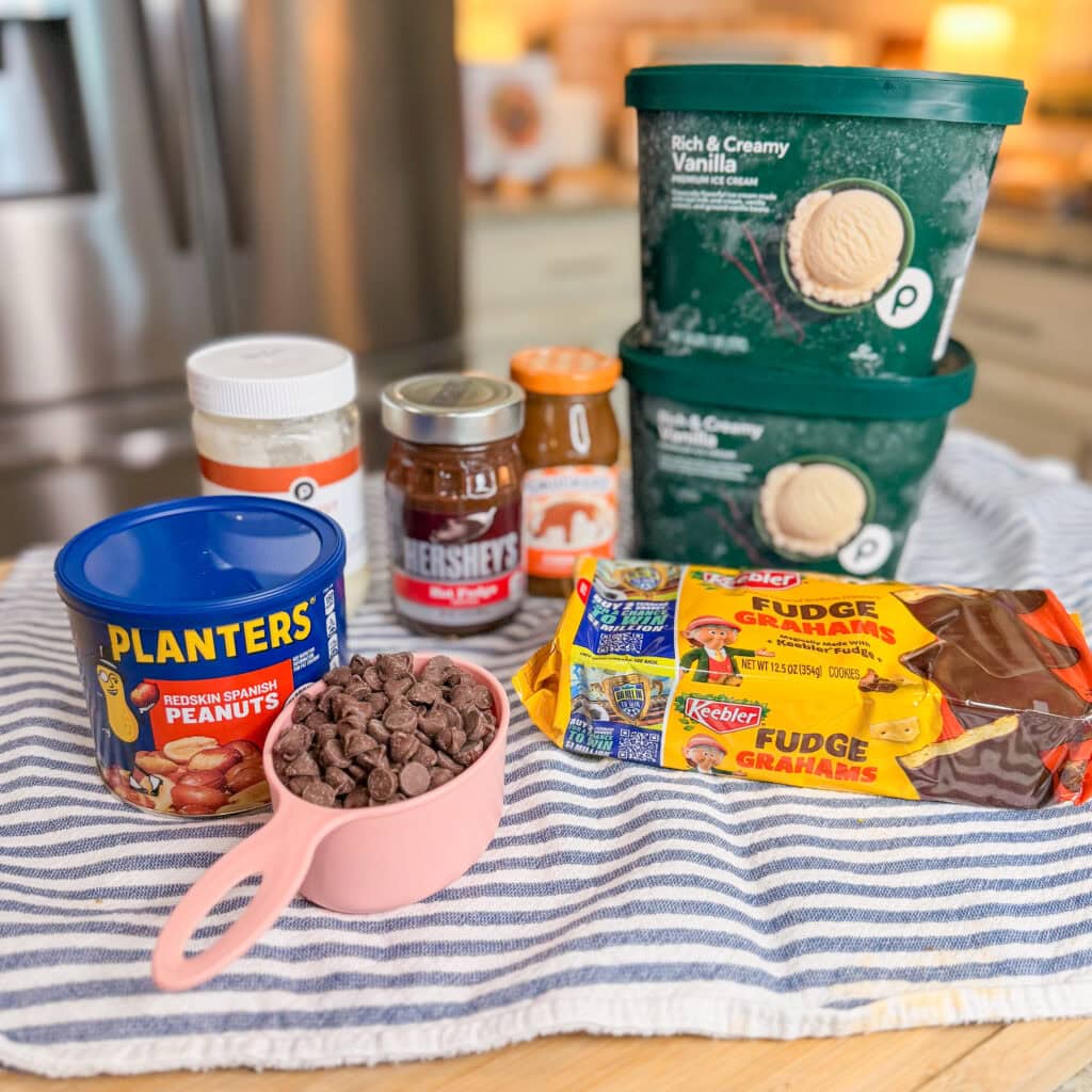 A striped cloth displays vanilla ice cream tubs, a can of peanuts, a jar of peanut butter, chocolate syrup, a jar of preserves, a cup of chocolate chips, and two packs of fudge graham crackers on a kitchen counter.