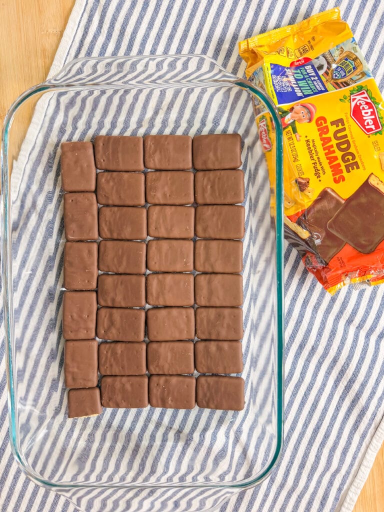 A glass baking dish with chocolate-covered fudge graham cookies neatly lined up on the bottom. Next to the dish are two packages of Keebler Fudge Grahams on a blue and white striped cloth.