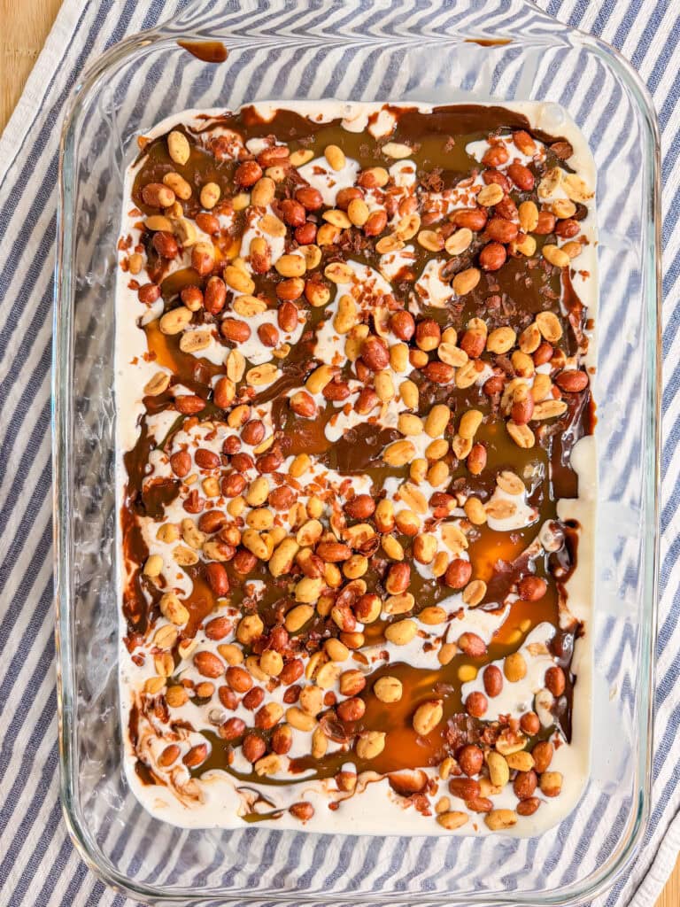 A glass baking dish filled with a dessert topped with roasted peanuts, caramel, and chocolate sauce, resting on a white and blue striped cloth.