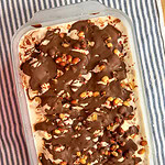 A rectangular dish of ice cream topped with drizzled chocolate and chopped nuts sits on a blue and white striped cloth.