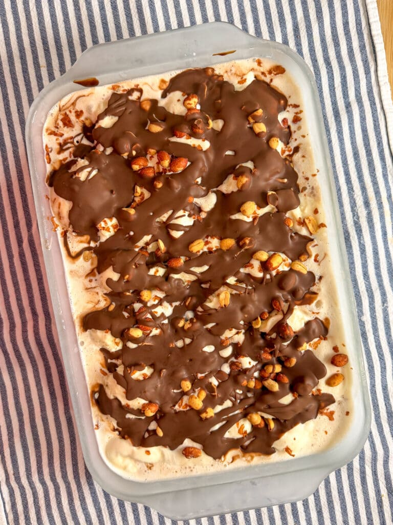 A rectangular dish of ice cream topped with drizzled chocolate and chopped nuts sits on a blue and white striped cloth.