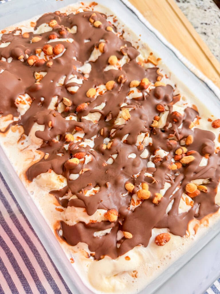 A glass dish filled with ice cream topped with a drizzle of chocolate and sprinkled with chopped nuts, placed on a striped cloth on a countertop.