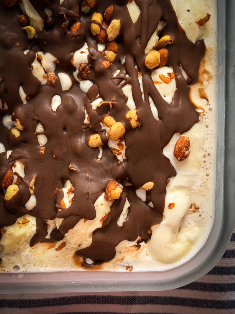 Close-up of vanilla ice cream topped with a hard chocolate shell and sprinkled with roasted peanuts in a glass container. The ice cream is melting slightly around the edges.