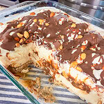 A glass baking dish filled with a layered dessert featuring a creamy filling, chocolate drizzle, and chopped nuts on top, with a portion removed to show the inside. The dish rests on a blue and white striped cloth.