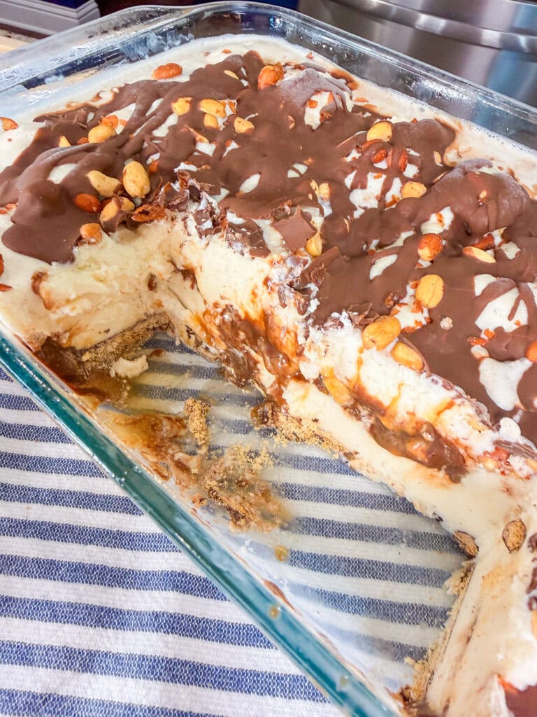 A glass baking dish filled with a layered dessert featuring a creamy filling, chocolate drizzle, and chopped nuts on top, with a portion removed to show the inside. The dish rests on a blue and white striped cloth.