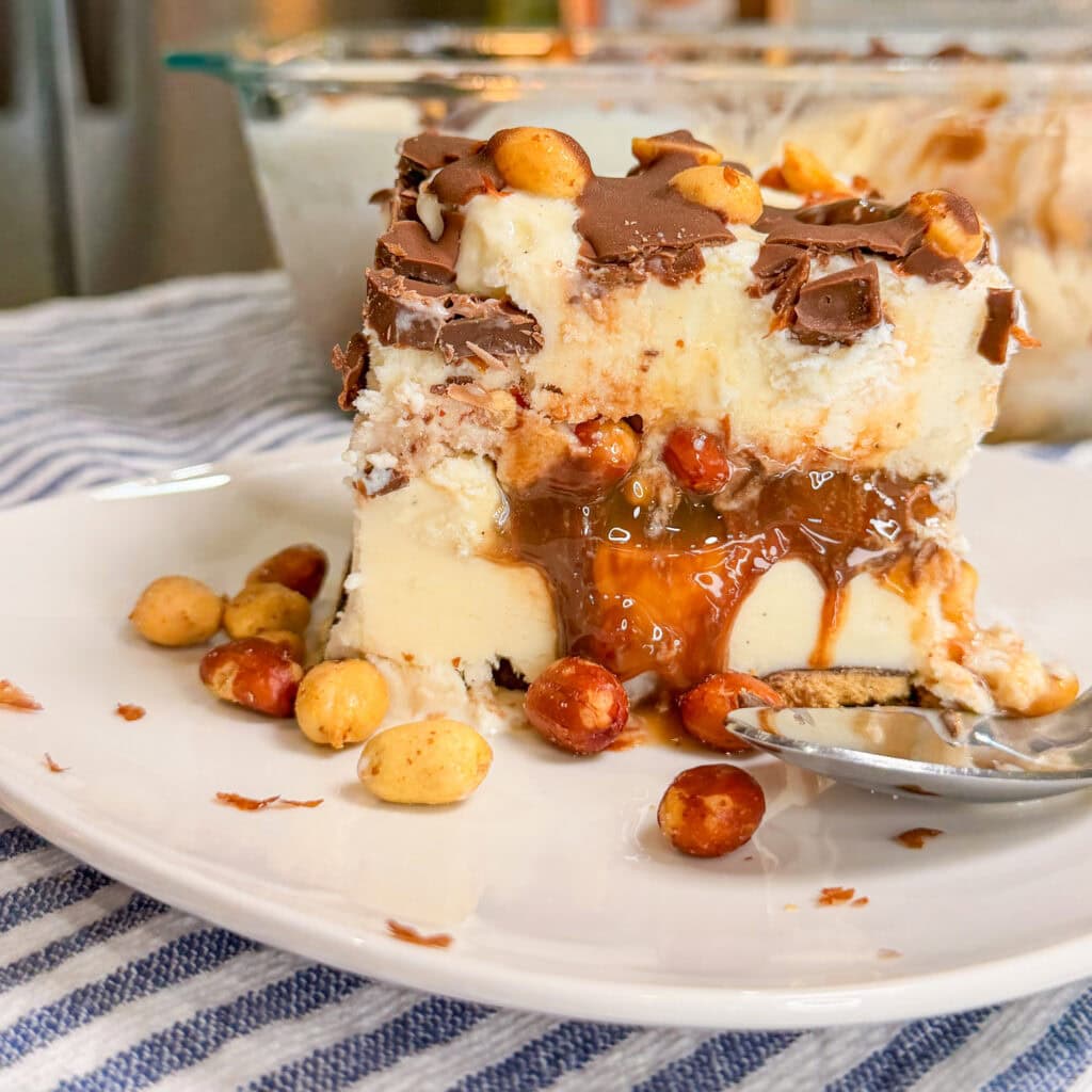 A close-up of a rich ice cream dessert slice topped with chocolate, caramel, and peanuts, served on a white plate with a spoon and striped cloth underneath.