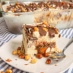 A slice of layered ice cream dessert topped with chocolate, caramel, and peanuts sits on a white plate with a spoon, with more peanuts scattered around and a larger dish of the dessert in the background.