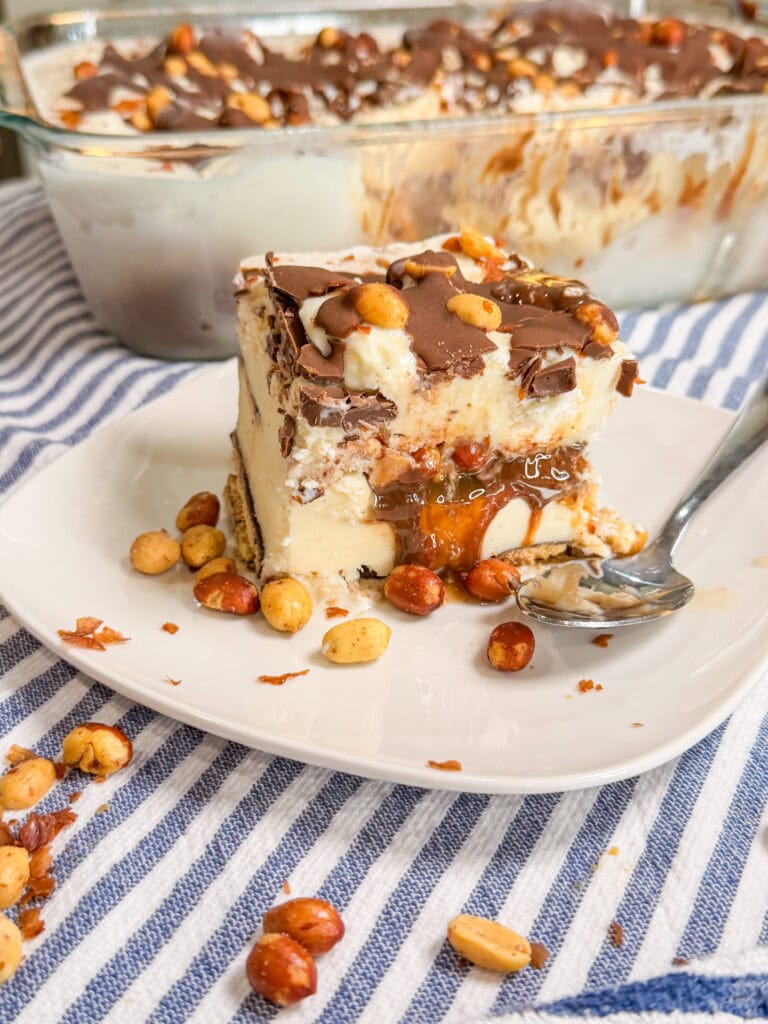 A slice of layered ice cream dessert topped with chocolate, caramel, and peanuts sits on a white plate with a spoon, with more peanuts scattered around and a larger dish of the dessert in the background.