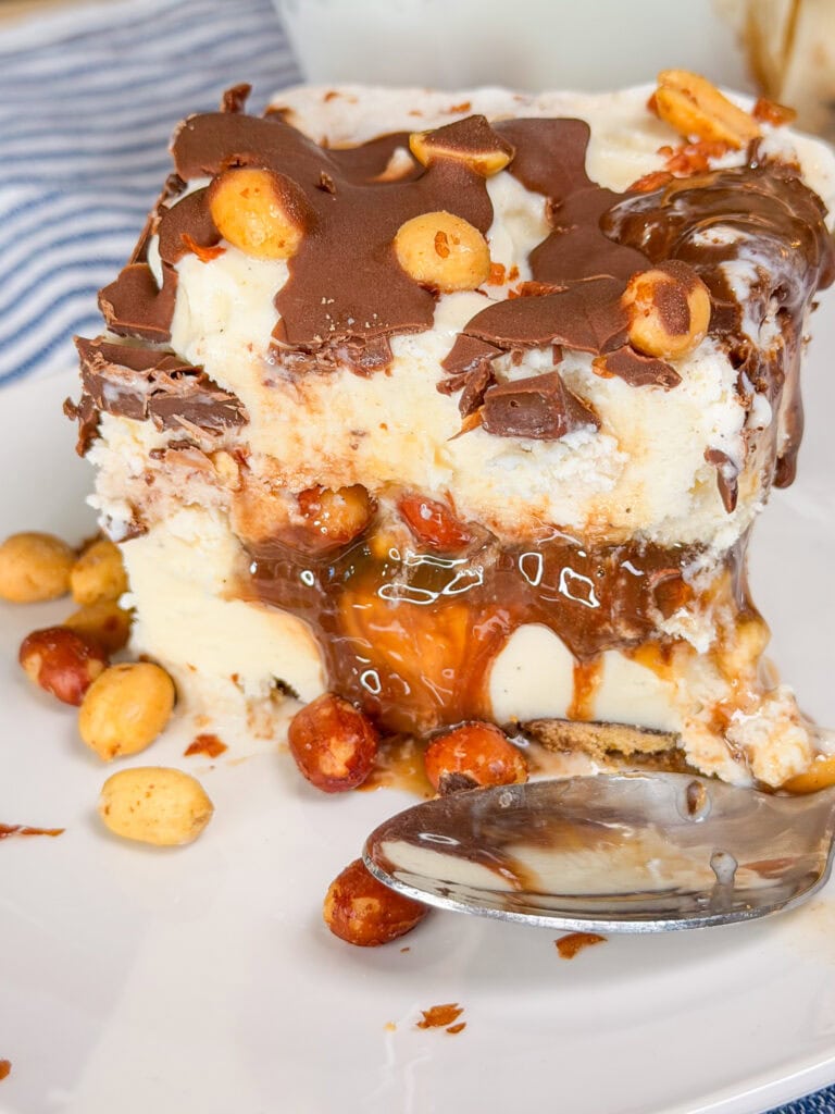 A slice of ice cream cake with chocolate shell, caramel, peanuts, and a cookie base sits on a white plate next to a spoon. Some peanuts and caramel sauce are scattered around the dessert.