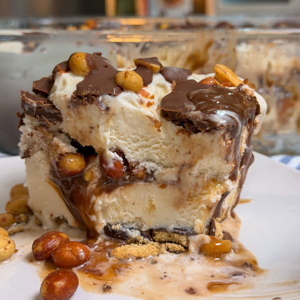 A close-up of a slice of ice cream dessert with layers of vanilla ice cream, chocolate topping, caramel, crushed cookies, and roasted peanuts, served on a white plate with a glass dish of the same dessert in the background.