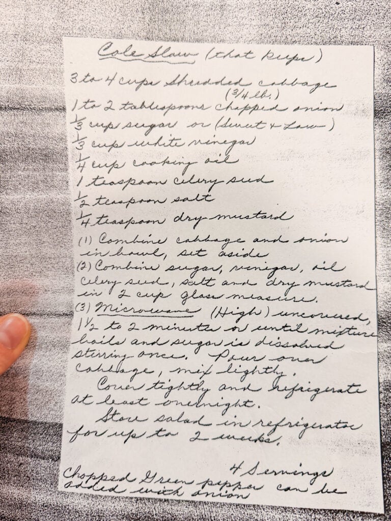 A handwritten recipe for coleslaw on white paper. Ingredients like shredded cabbage, chopped onion, sugar, and vinegar are listed, with instructions for mixing and microwaving. A persons thumb holds the page on the left.