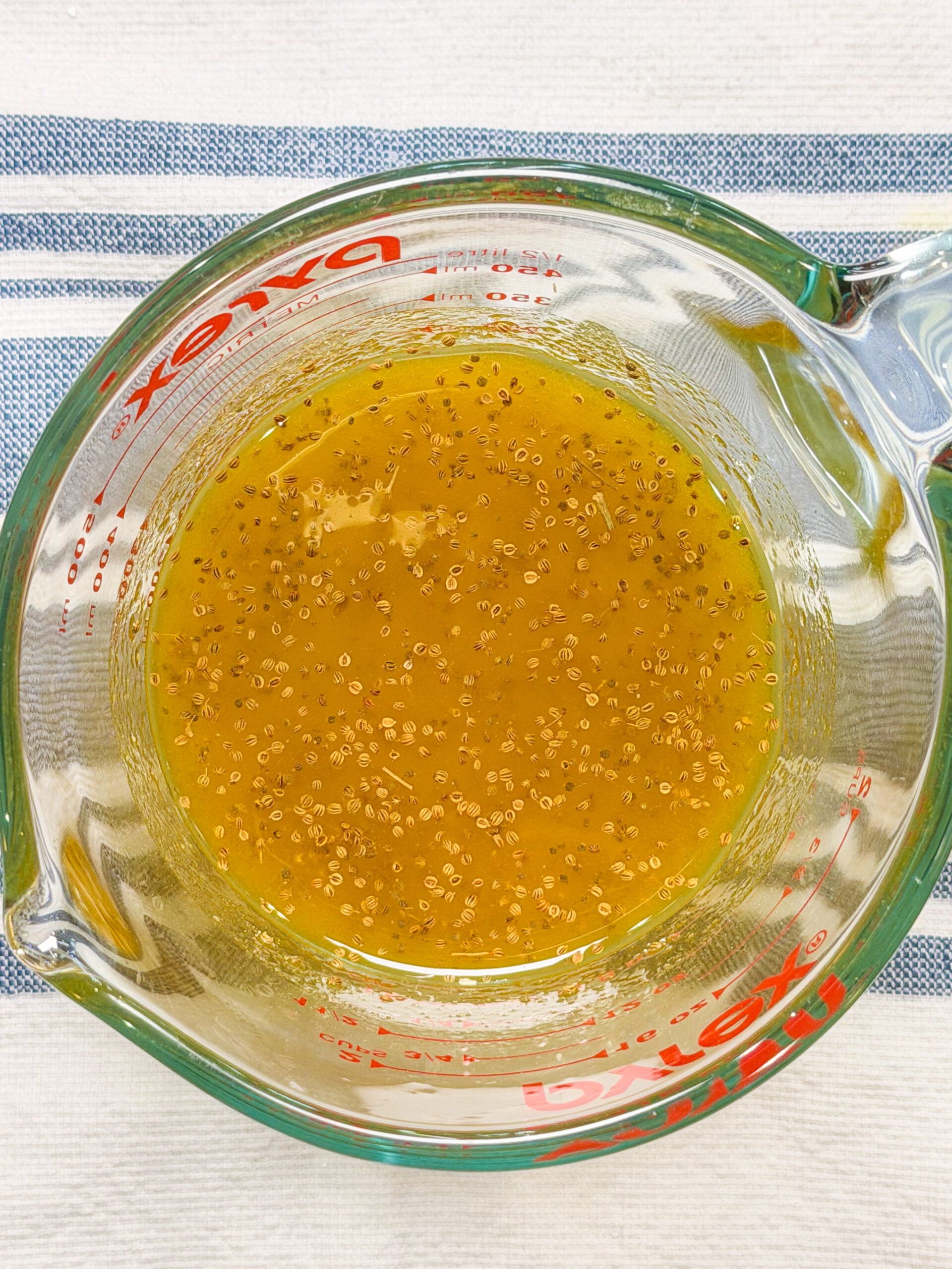 A glass Pyrex measuring cup filled with a yellow-orange liquid containing small chia seeds, placed on a white cloth with blue stripes.