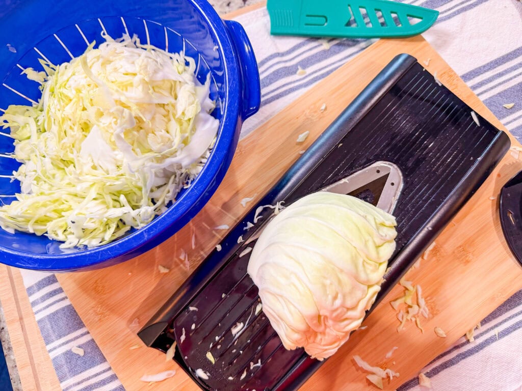A half head of cabbage sits on a mandoline slicer on a wooden cutting board, with shredded cabbage collected in a blue colander nearby. A green vegetable peeler is also on the table.