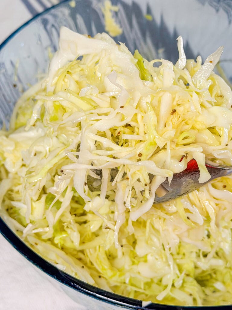 A close-up of shredded cabbage salad in a glass bowl, lightly dressed and being mixed with a metal spoon.