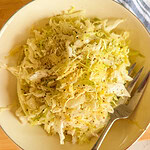 A bowl of shredded cabbage salad topped with black pepper, with a fork resting on the side of the bowl. The dish sits on a light surface with part of a blue-striped cloth visible.