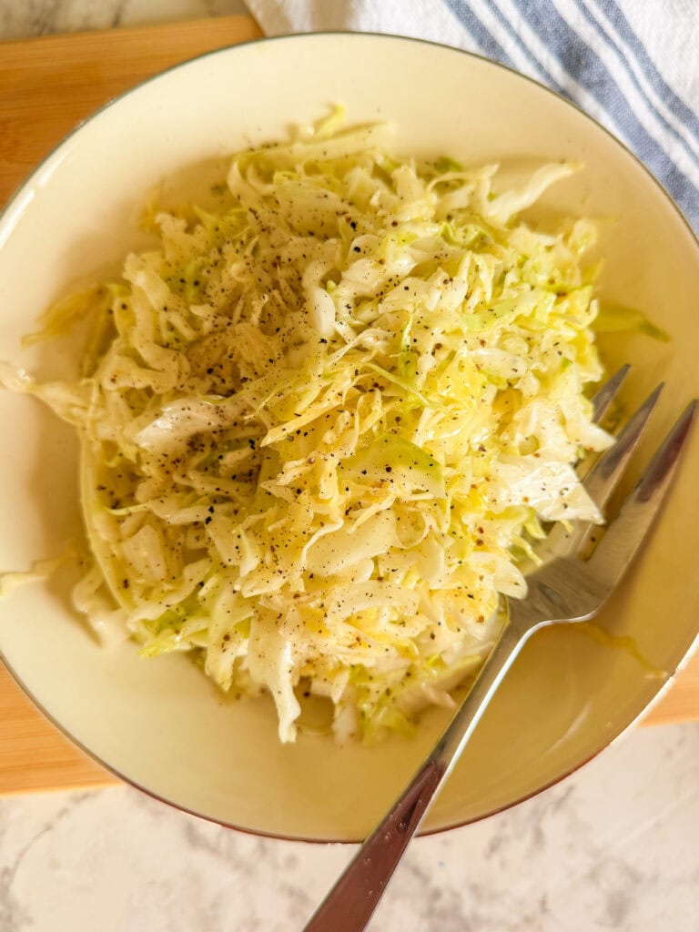 A bowl of shredded cabbage salad topped with black pepper, with a fork resting on the side of the bowl. The dish sits on a light surface with part of a blue-striped cloth visible.