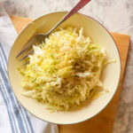 A bowl of shredded cabbage salad sprinkled with black pepper, placed on a wooden board with a metal fork and a white towel with blue stripes nearby.