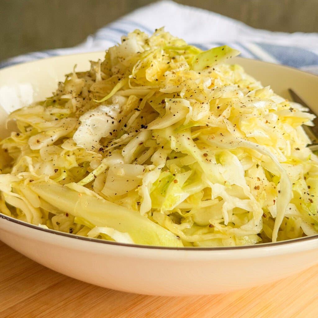 A close-up of a bowl filled with shredded cabbage salad, lightly seasoned with black pepper, set on a wooden surface with a blue and white cloth in the background.