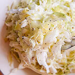 A close-up of a bowl of shredded cabbage salad, lightly seasoned with black pepper, on a white plate. A spoon is partially visible in the salad.