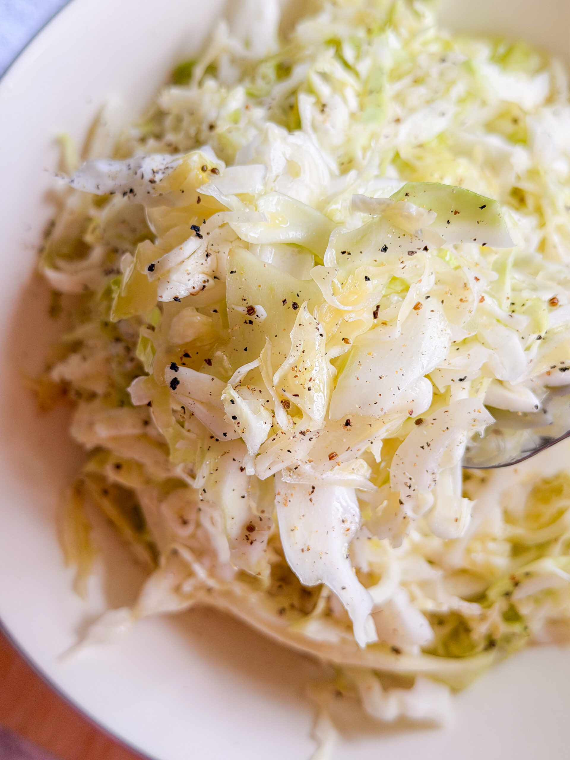 A close-up of a bowl of shredded cabbage salad, lightly seasoned with black pepper, on a white plate. A spoon is partially visible in the salad.