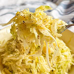 A close-up of a fork holding a bite of creamy coleslaw, with shredded cabbage dressed in mayonnaise and sprinkled with black pepper, above a bowl filled with more coleslaw.