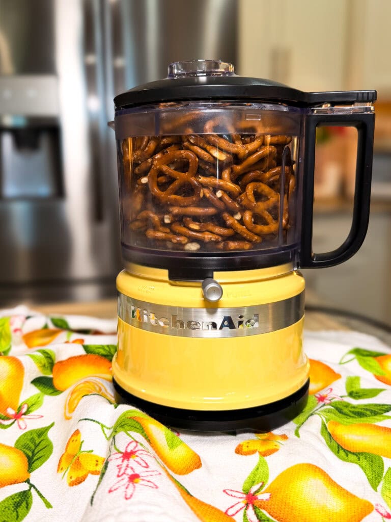 A yellow KitchenAid food processor filled with pretzels sits on a lemon-patterned cloth in a kitchen, with a refrigerator and cabinets visible in the background.