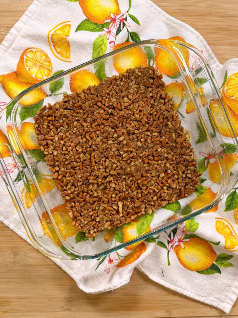 A glass baking dish with a baked pecan-topped dessert sits on a lemon-patterned kitchen towel atop a wooden surface.