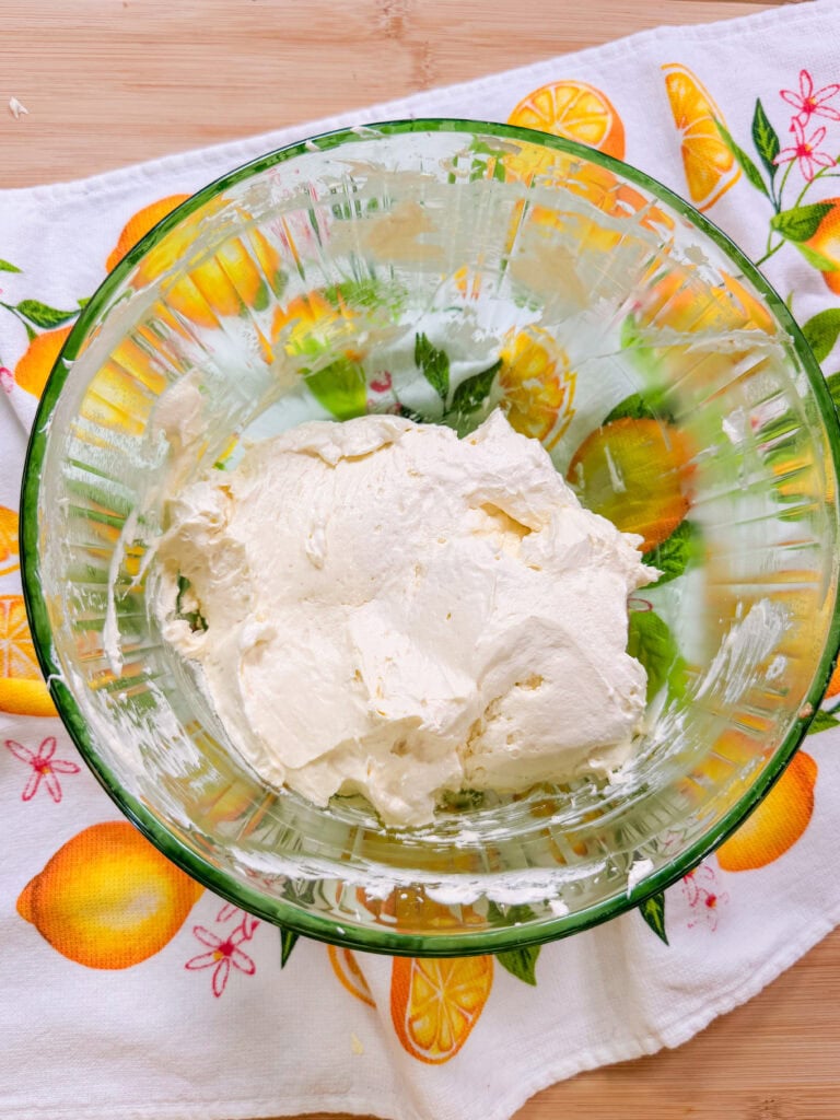 A glass bowl filled with a mound of creamy white cheese sits on a white cloth decorated with colorful lemons and orange slices. The background is a light wooden surface.