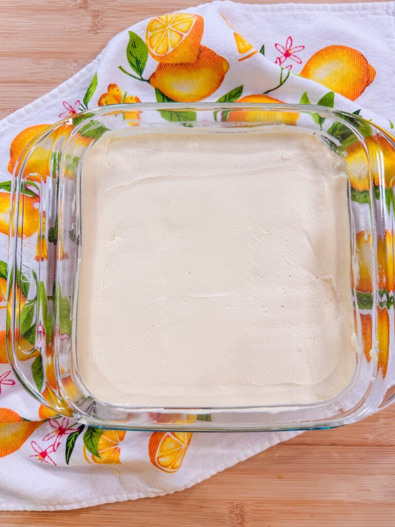 A glass baking dish filled with a smooth, creamy white mixture sits on a lemon-patterned towel on a wooden surface.