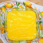 A glass baking dish filled with smooth, bright yellow lemon filling sits on a lemon-patterned towel atop a wooden surface.