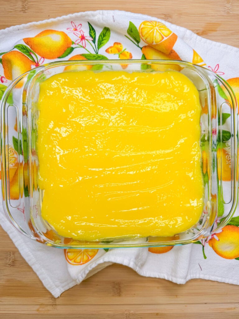 A glass baking dish filled with smooth, bright yellow lemon filling sits on a lemon-patterned towel atop a wooden surface.