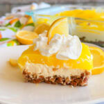 A slice of lemon dessert with a pretzel crust, creamy filling, and bright yellow lemon topping, garnished with whipped cream and a lemon slice, sits on a white plate with lemon slices in the background.