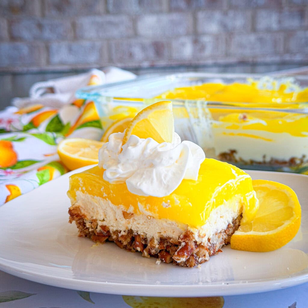 A slice of layered lemon dessert with a nutty crust, creamy middle, and bright yellow lemon topping, garnished with whipped cream and a lemon slice, sits on a white plate with lemon halves in the background.