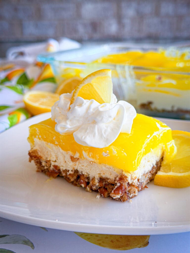 A slice of lemon dessert with a nutty crust, creamy middle layer, and bright yellow lemon topping, garnished with whipped cream and a lemon wedge on a white plate. A baking dish with more dessert is in the background.