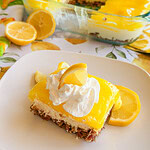 A square slice of lemon dessert with a crumb crust, creamy middle layer, and bright yellow lemon topping, garnished with whipped cream and lemon slices on a white plate. The rest of the dessert is in a glass baking dish.