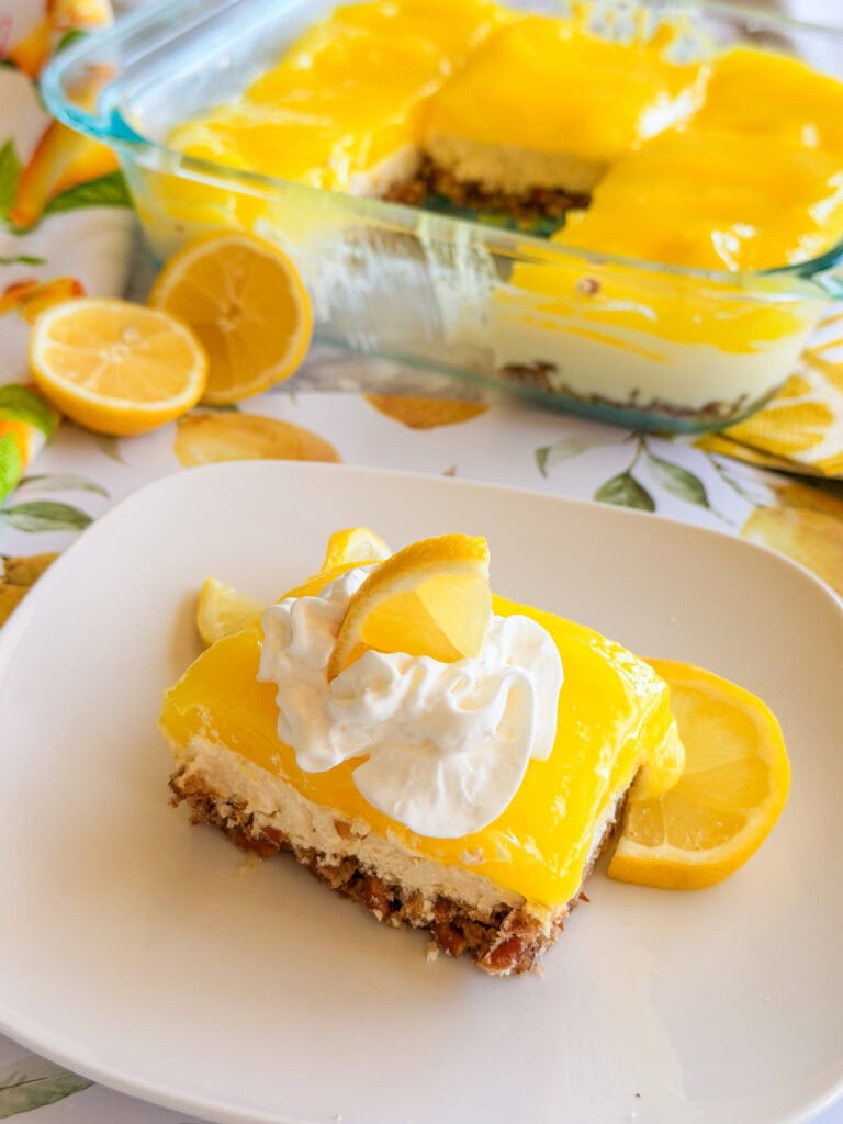A square slice of lemon dessert with a crumb crust, creamy middle layer, and bright yellow lemon topping, garnished with whipped cream and lemon slices on a white plate. The rest of the dessert is in a glass baking dish.