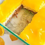 A glass baking dish with lemon bars, topped with bright yellow lemon curd, one section missing to reveal a crumbly crust and creamy filling. The dish is on a surface with a lemon pattern.