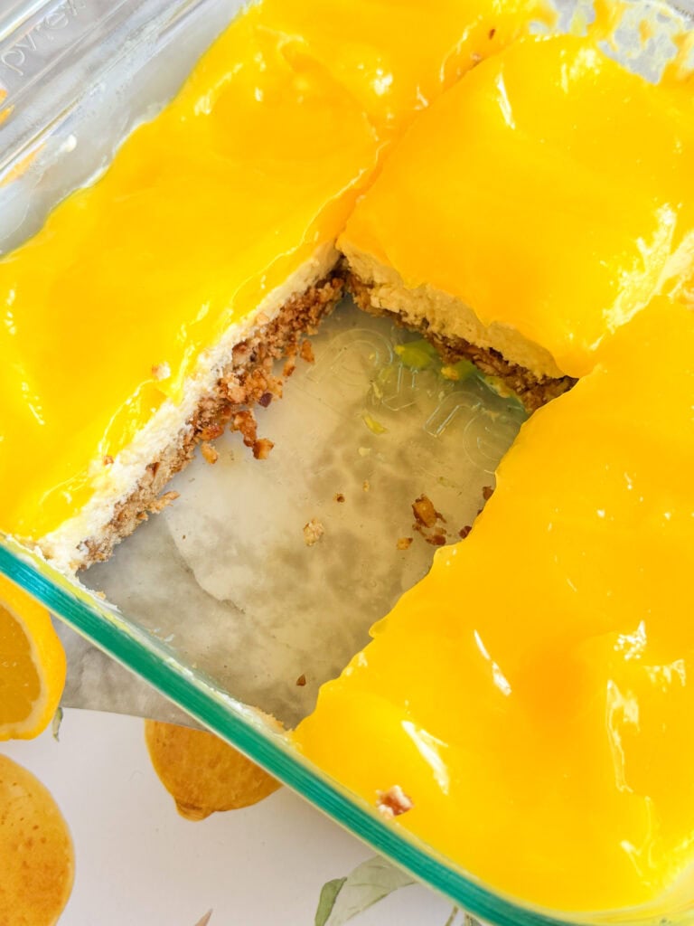 A glass baking dish with lemon bars, topped with bright yellow lemon curd, one section missing to reveal a crumbly crust and creamy filling. The dish is on a surface with a lemon pattern.