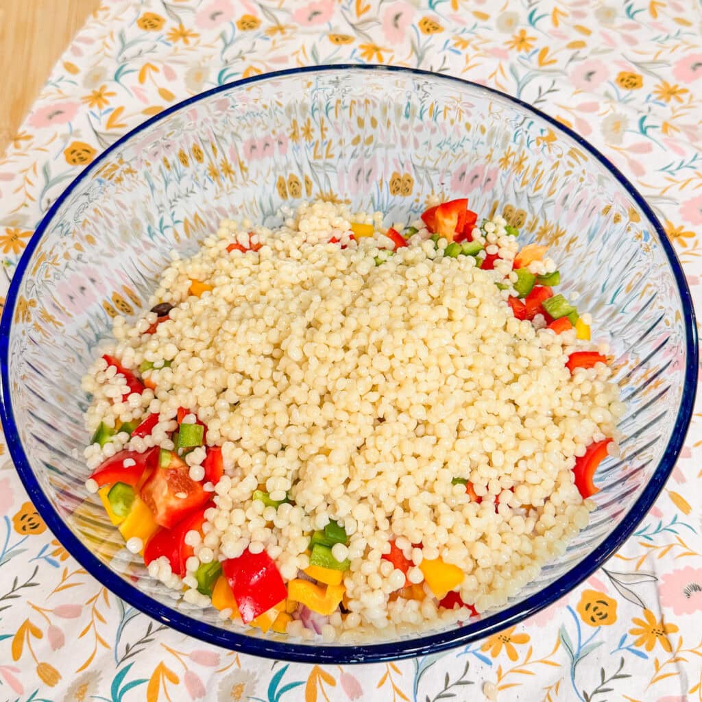 A glass bowl filled with pearl couscous mixed with chopped red, yellow, and green bell peppers sits on a floral-patterned tablecloth.